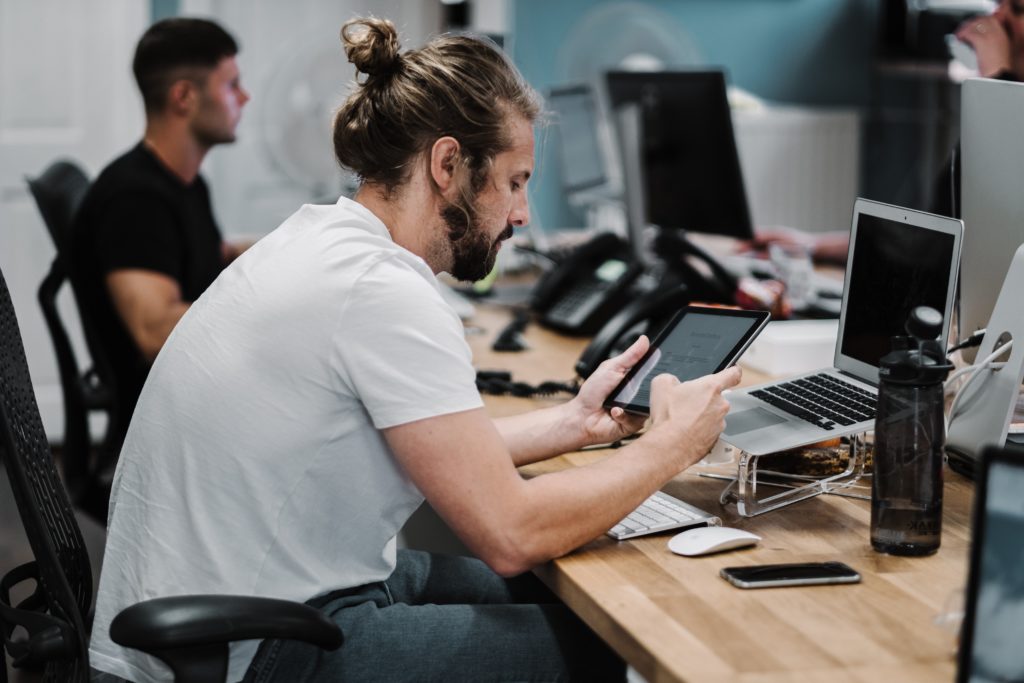 Foto von einem Mann mit langen Haaren, der in einem IT-Büro sitzt und konzentriert auf einem Tablet herumtippt.
